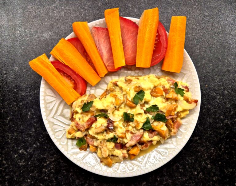 Jajecznica z kurkami i boczkiem podana na talerzu z marchewką i pomidorem.