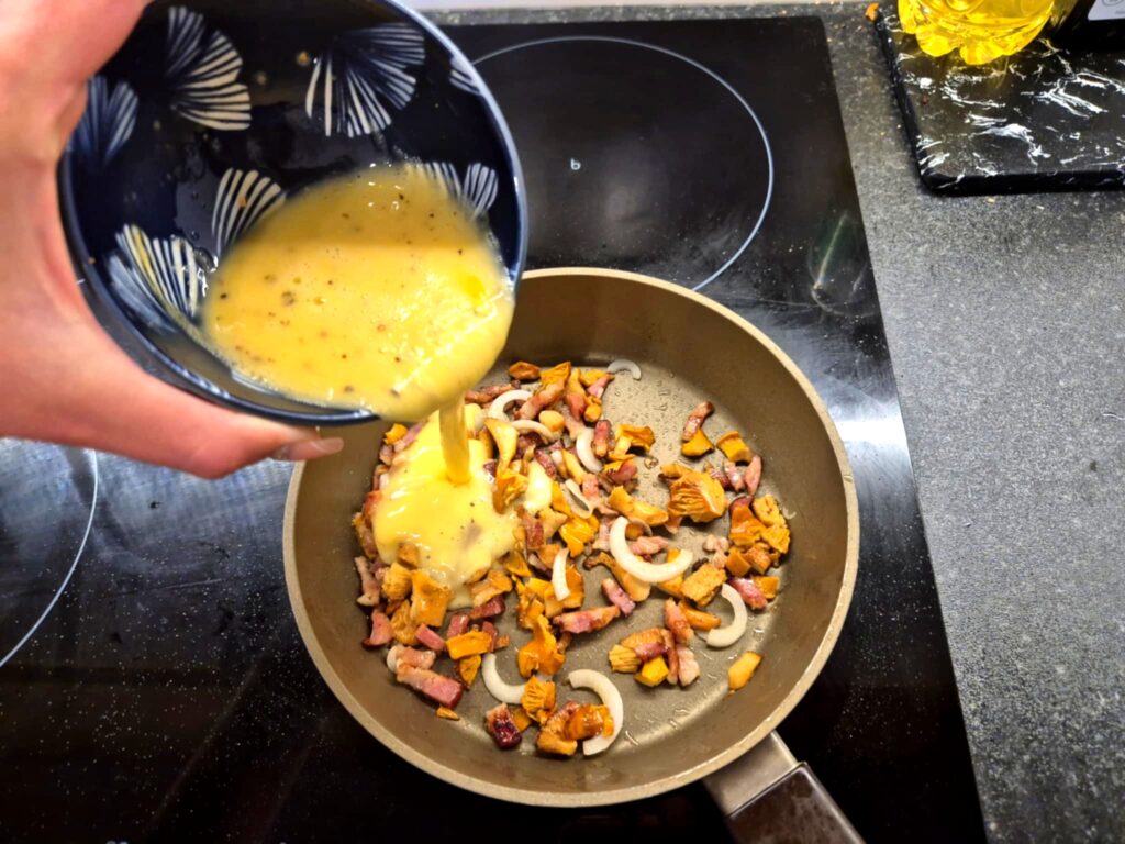 Roztrzepane jajka wlewane na patelnię z boczkiem i kurkami.